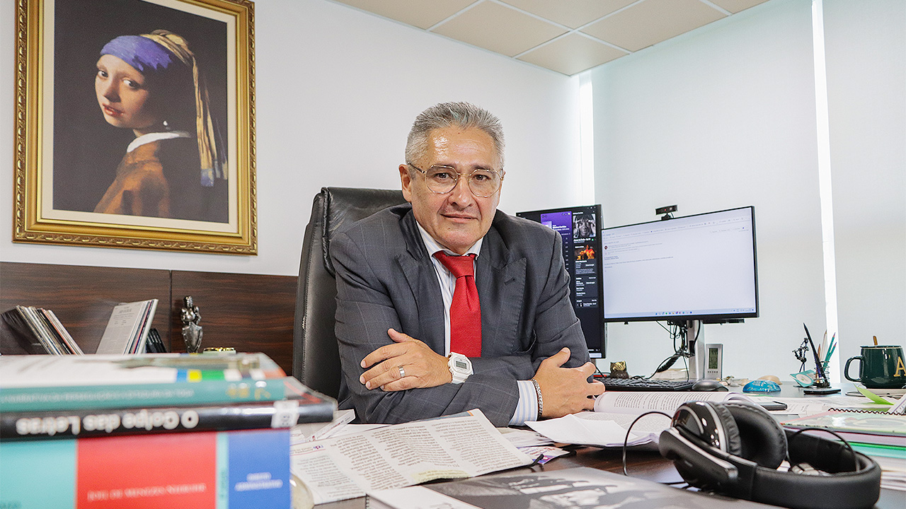 Foto do conselheiro Aderson Flores em sua sala. Ele está sentado diante da sua mesa, sobre a qual há livros e papéis. Aderson é um homem branco, de cabelos grisalhos. Veste terno cinza-escuro e gravata vermelha. Está sorrindo, com os braços apoiados sobre a mesa. Ao fundo, na parede, há a pintura “Moça com brinco de pérola”, do artista Johannes Vermeer. 