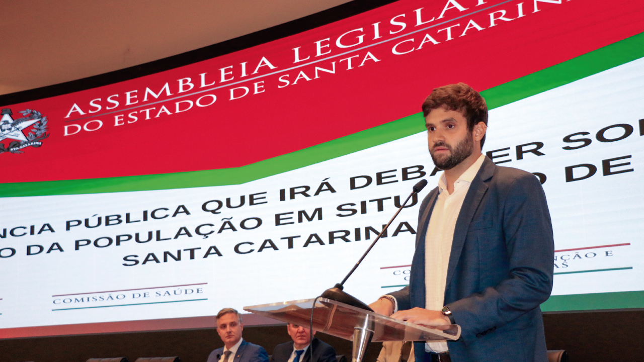 Foto do auditor fiscal Rafael Scherb, em frente ao púlpito, falando ao microfone. Ao fundo, painel luminoso com o banner identificando o nome do evento, e dois deputados sentados. 