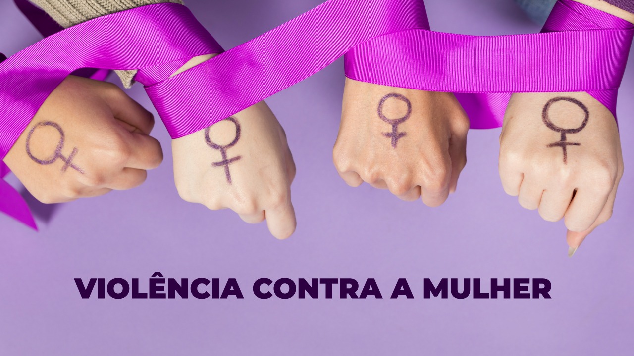 Foto de quatro punhos fechados e virados para baixo, um ao lado do outro. As mãos são de mulheres brancas. Sobre o dorso de cada uma delas, há o desenho do símbolo feminino. Uma fita rosa passa por cima e se enrola nas mãos. O fundo é lilás. Na parte inferior, há o texto “Violência contra a mulher”, em fonte roxa.