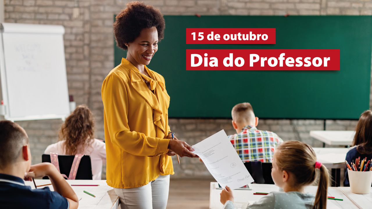 Foto de uma professora entregando uma folha de papel a uma criança sentada em uma carteira. A professora é uma mulher negra, de cabelos crespos, que sorri e usa roupa amarela. Há outras crianças sentadas em carteiras enfileiradas em uma sala de aula. Ao fundo, no quadro de giz, o texto “15 de outubro. Dia do Professor”, em destaque em retângulos vermelhos.