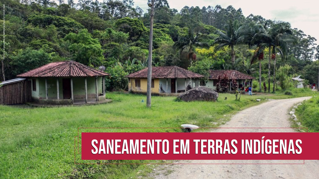 Imagem mostra a aldeia guarani em Canelinha. A foto mostra três casas em um gramado verde, com mata aos fundos e uma estrada sem pavimentação à direita. Na parte inferior da imagem há uma tarja vermelha com a inscrição “Saneamento em terras indígenas” em branco