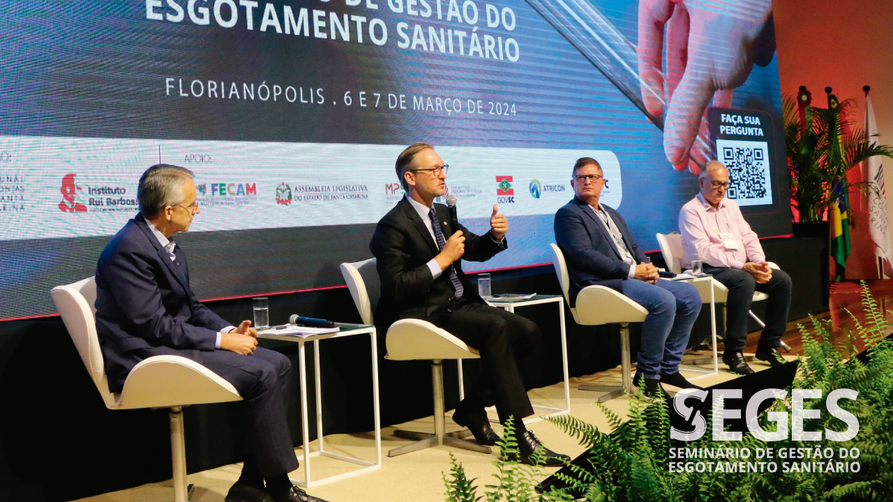 Foto dos quatro participantes do painel durante o Seminário. Da esquerda para a direita, o presidente da Agir, Mário Hildebrandt, o desembargador do TJSC Marcos Fey Probst, o presidente da Aresc, João Carlos Grando, e o presidente, em 2023, do Conselho de Administração da Aris, Mario Afonso Woitexem. Ambos estão sentados em poltronas, no palco do auditório do evento, enquanto debatem. Ao fundo, projeção do logo do Seges no telão. 