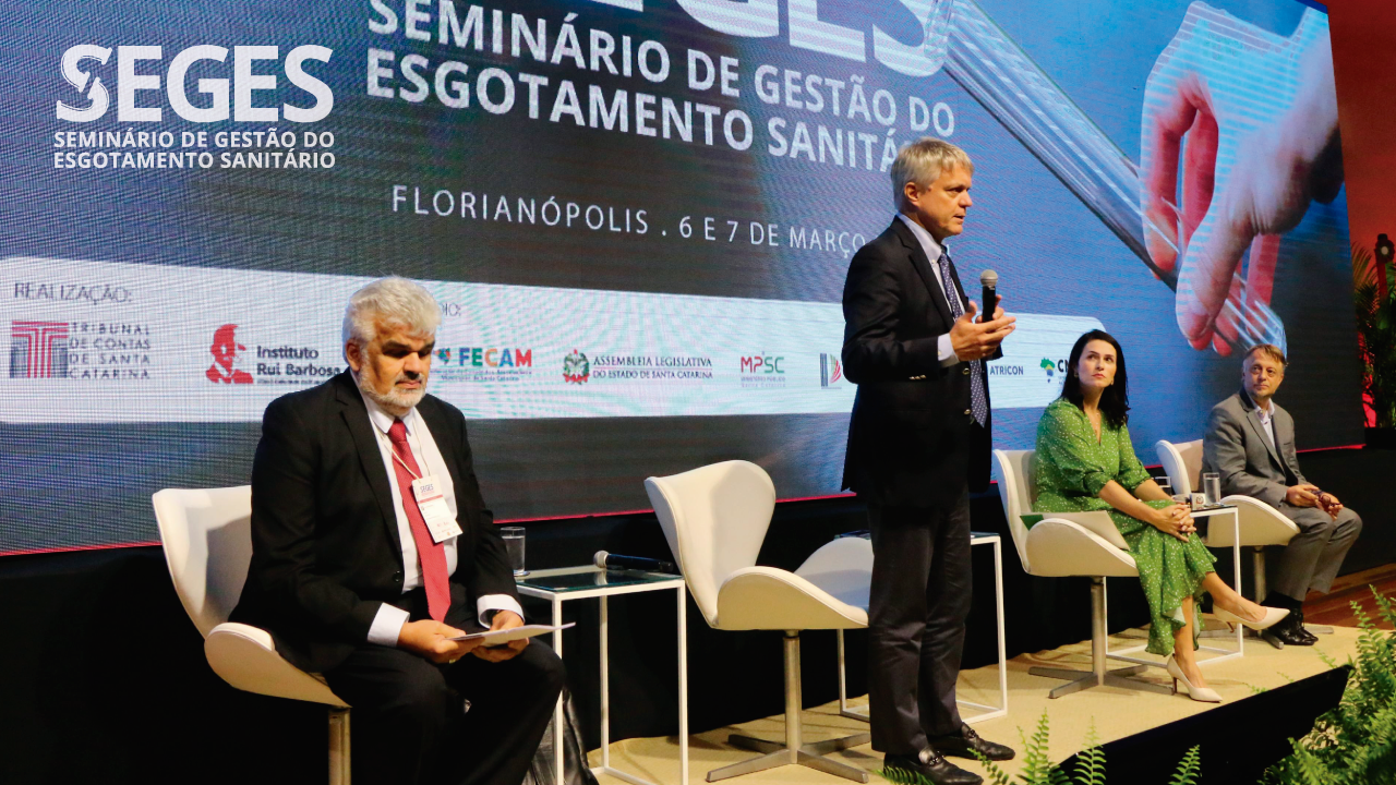 Foto dos quatro participantes do painel durante o Seminário. Da esquerda para a direita, o presidente do Comitê Técnico do Meio Ambiente e Sustentabilidade do IRB, conselheiro Júlio Pinheiro, o presidente do CNPTC, conselheiro Luiz Antonio Guaraná, a coordenadora do Centro de Apoio Operacional do Meio Ambiente, promotora de Justiça do MPSC Fernanda Broering Dutra e o procurador-geral do MPTC/SC, Diogo Ringenberg. Ao fundo, projeção do logo do Seges no telão. 