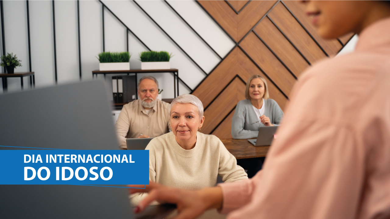 Banner horizontal com foto de três idosos sentados diante de mesas em uma sala, olhando para o que mostra uma mulher que está à frente deles, de costas para a foto, em primeiro plano e com desfoque. Sobre a imagem, na lateral esquerda, em tarja azul, o texto “Dia Internacional do Idoso”.