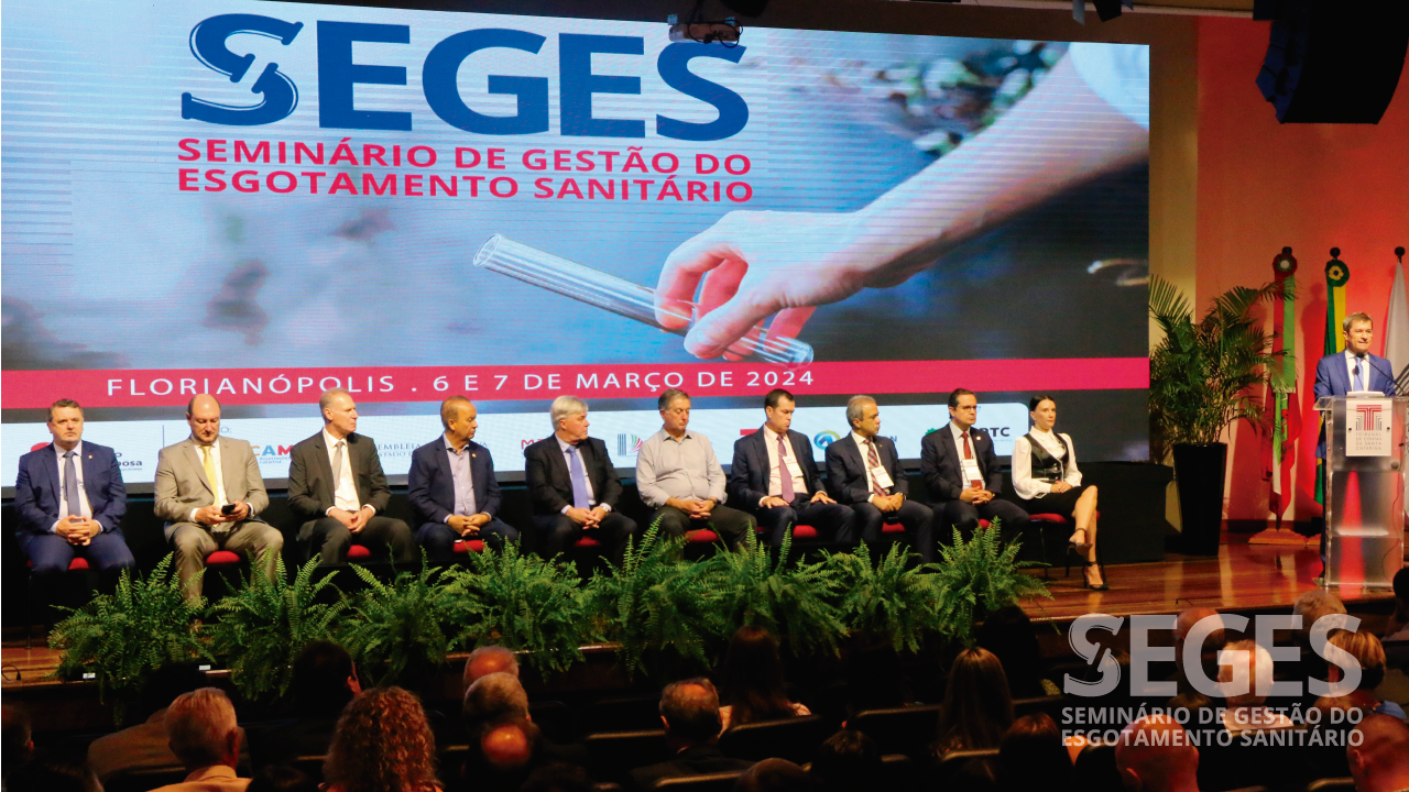 Foto das autoridades no palco do auditório da Alesc, durante o evento. São 10 pessoas, sentadas em cadeiras. No púlpito, fala o vice-presidente, conselheiro José Nei Ascari. Ao fundo, no telão, o logo do Seminário. 