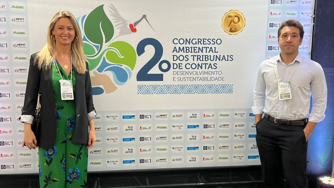 Foto horizontal dos auditores fiscais de controle externo Ana Sophia Besen Hillesheim (à esquerda) e Jean Rodrigues de Souza (à direita) em frente ao banner do evento. 