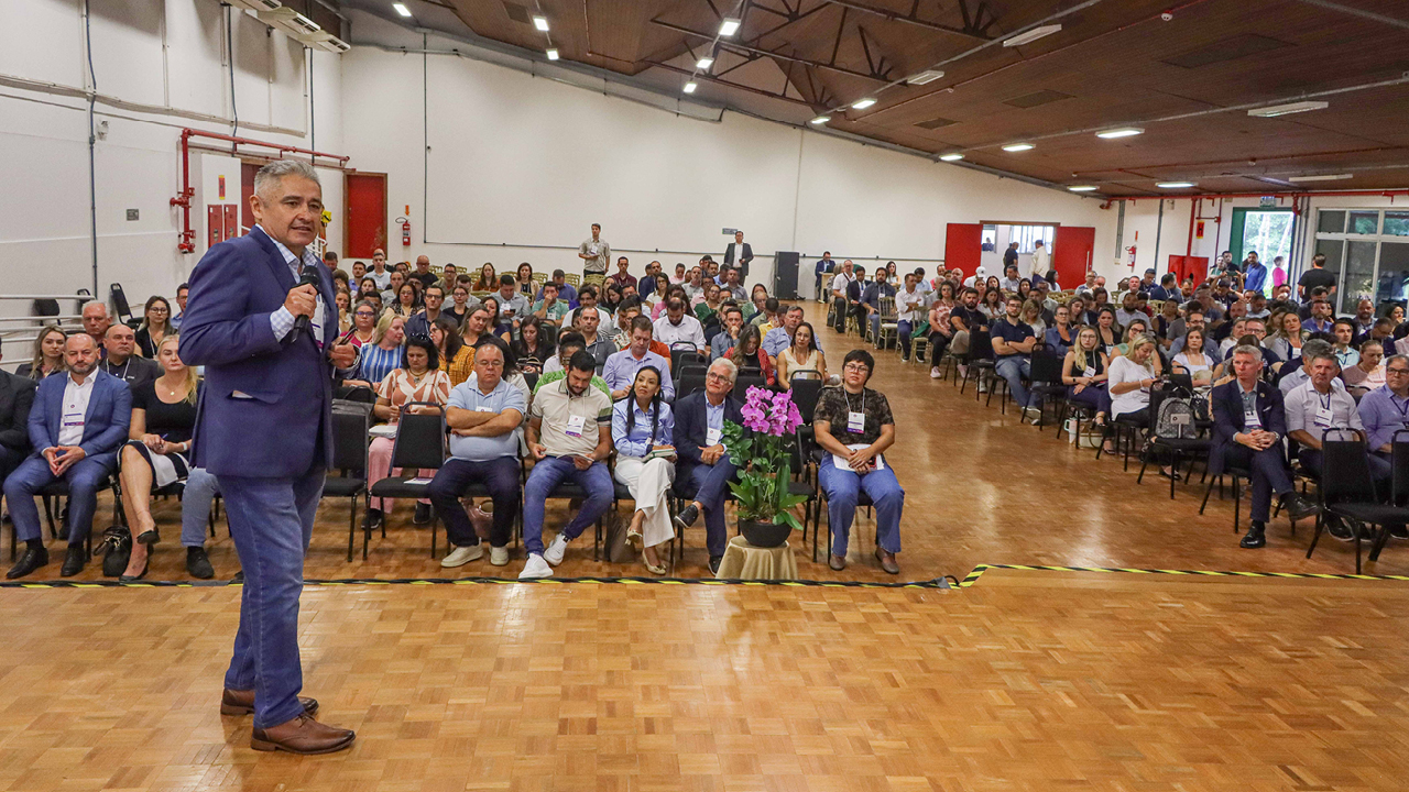 Foto mostra um auditório cheio ao fundo.  À esquerda, de frente para a câmera, está um homem de traje escuro. 