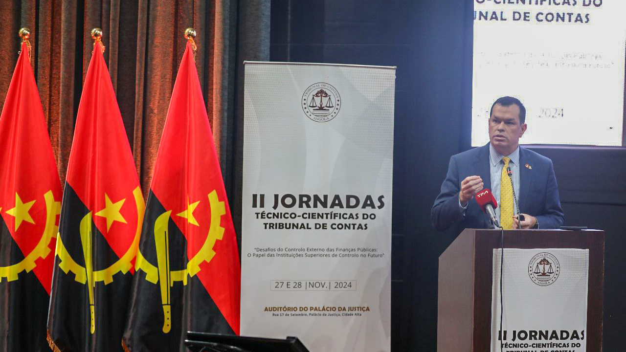 Foto do conselheiro Adircélio de Moraes palestrando. Ele está atrás do púlpito, à direita, e veste terno azul marinho. Ao fundo do palco e a frente do púlpito, o banner do evento. À esquerda da foto, três bandeiras de Angola hasteadas.  