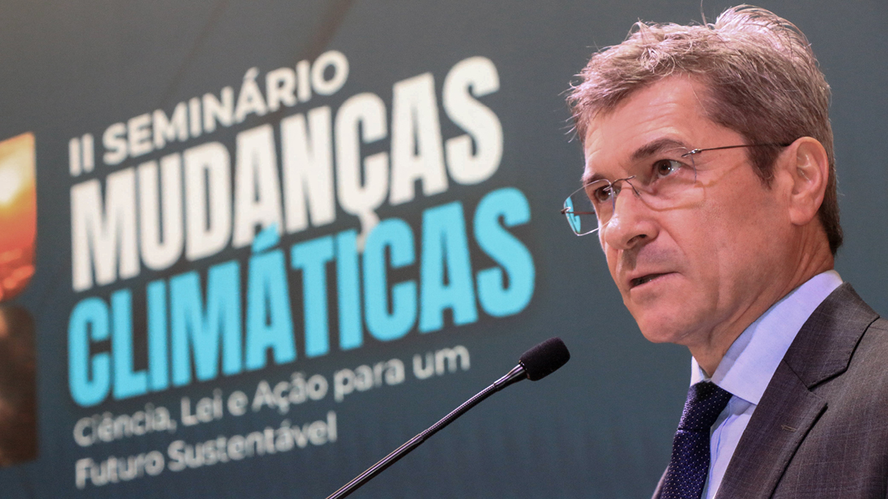 Foto do conselheiro José Nei Ascari discursando no púlpito. Ele é um homem branco, de cabelos grisalhos. Veste terno e gravata e usa óculos de grau. Ao fundo, banner com o título do evento, “segundo Seminário Mudanças Climáticas: ciência, lei e ação para um futuro sustentável”.