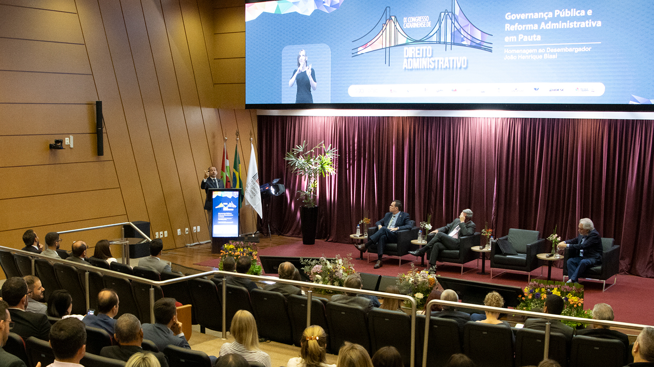 Banner horizontal com foto que mostra parte da plateia, de costas, assistindo a um dos painéis do congresso. Há várias pessoas sentadas em poltronas do auditório, lado a lado. No palco, entre os presentes, os desembargadores Hélio Pereira e João Henrique Blasi, o conselheiro Adircélio Ferreira Júnior e o presidente do Idasc, José Cristóvam. Ao fundo, no telão, é exibida a identidade visual do evento.  