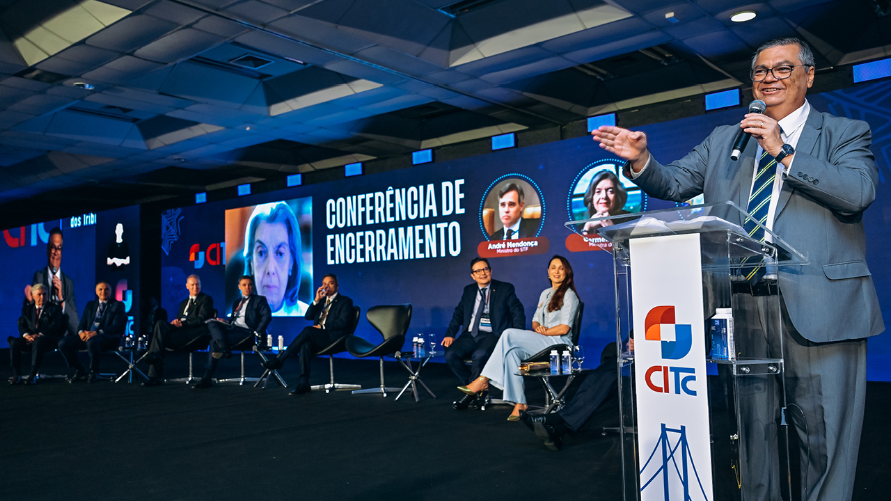 Foto do palco do IV CITC. À direita, no púlpito, o ministro do STF Flavio Dino discursa. Ao funo, telão com imgame da ministra Carmen Lucioa e os dizeres Conferencia de Encerramento. No palco ainda estão os presidentes do IRB, Atricon , TCE/SC r o ministro André Mendonça.