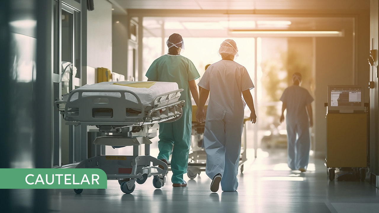 Banner horizontal. Imagem de um corredor de hospital. Nela, uma maca vazia à esquerda. Ao centro, de costas, dois enfermeiros caminham em direção à uma porta de vidro. No canto inferior direito, sobre um fundo verde claro, em letras brancas está escrita a palavra cautelar.