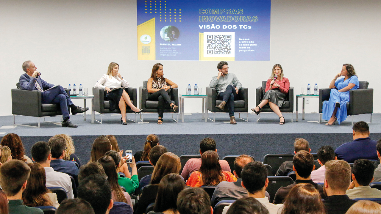 Foto do evento. Ao fundo, sobre o palco, o palestrante e os cinco debatedores, sendo quatro mulheres e um homem, sentados em poltronas pretas e de frente para a plateia, cujas pessoas também estão sentadas. 