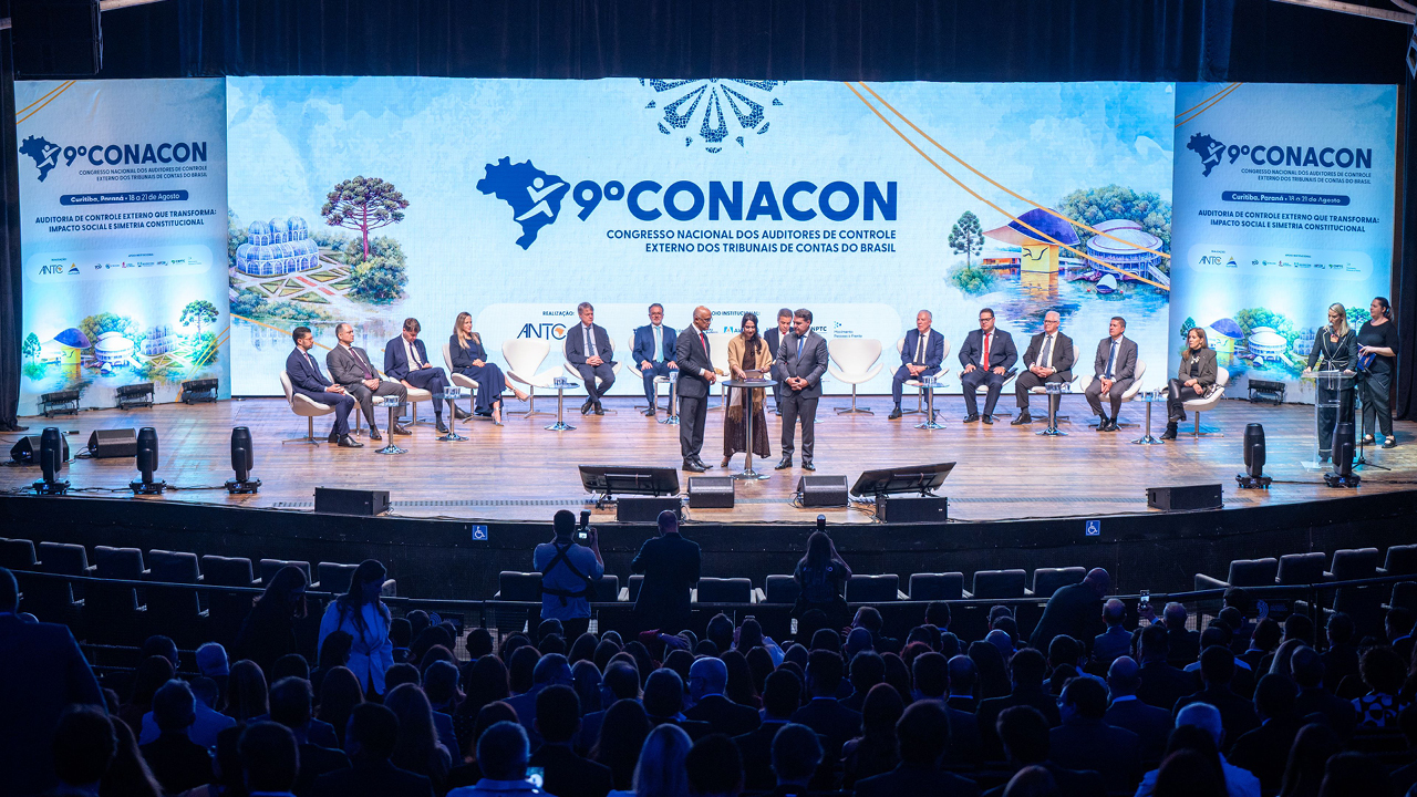 Banner horizontal com foto do auditório lotado durante a realização do 9º Conacon – Congresso Nacional dos Auditores de Controle Externo dos Tribunais de Contas do Brasil. No palco, autoridades e participantes estão distribuídos em cadeiras ao fundo, diante de um grande painel com a identidade visual do evento. Ao centro, a auditora fiscal de controle externo do TCE/SC, Juliana Moser, recebe uma homenagem durante a cerimônia, acompanhada por representantes da organização. 