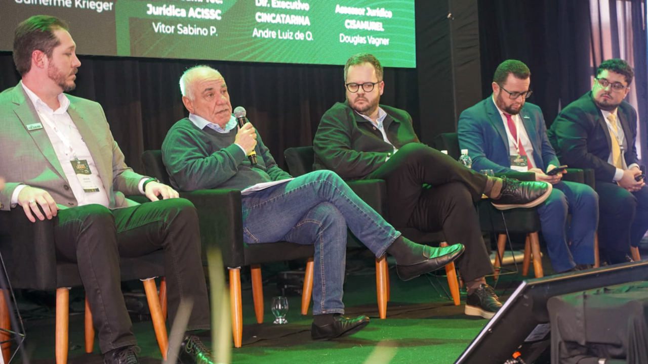 Imagem mostra cinco homens sentados em cadeiras em um palco. O segundo homem da esquerda para a direita, o conselheiro Luiz Eduardo Cherem, está falando ao microfone
