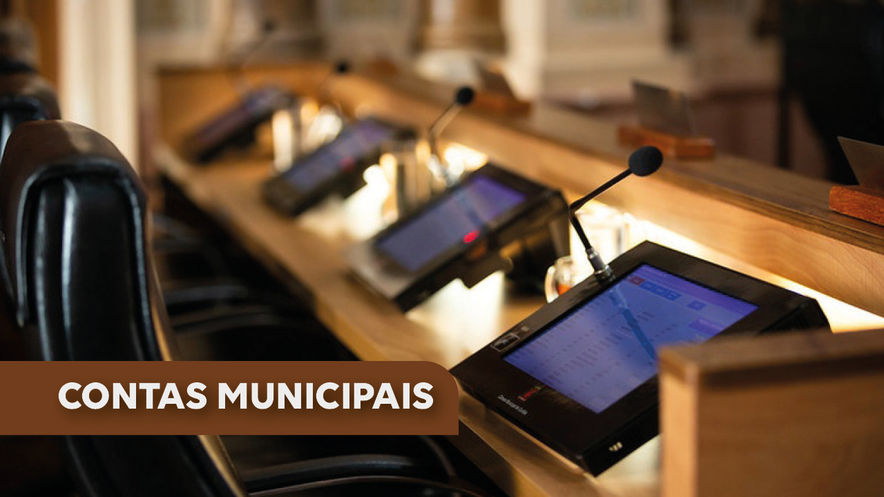   Imagem   Foto da mesa de uma bancada, em que aparecem quatro pequenos monitores em cujas telas aparece uma planilha com dados, e quatro microfones. A frente da bancada há cadeiras.  No canto inferior esquerdo há uma tarja na cor marrom, com o texto “Contas municipais” em fonte branca 