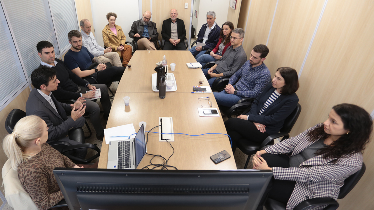 Foto de várias pessoas sentadas diante de uma mesa de reuniões durante apresentação dos novos módulos do SEI. Entre os presentes, o diretor de Tecnologia da Informação, Rafael Queiroz, que fala a outros integrantes do TCE/SC. Sobre a mesa, há notebook, papéis e celulares.