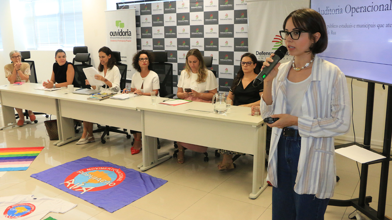 Foto do palco do evento. a auditora Sabrina Silva, uma mulher branca, com cabelos médios escuros está ao microfone. Atrás dela um painel eletrônico com a apresentação da sua palestra. Ao lado uma mesa retangular com seis mulheres.