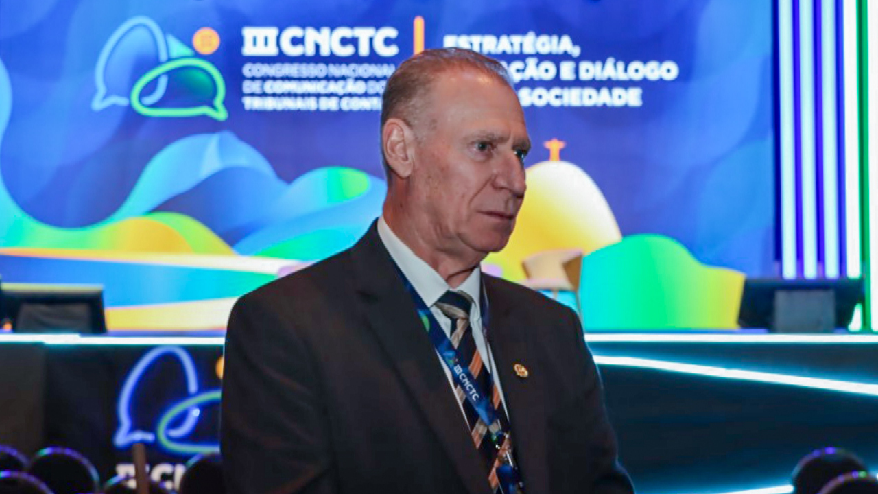 Foto do presidente do TCE/SC, conselheiro Herneus De Nadal, no III Congresso Naciional de Comunicação dos Tribunais de Contas. Eles estpa de terno escuro. Ao fundo, um painel em tons de azul e verso com a logo do evento,