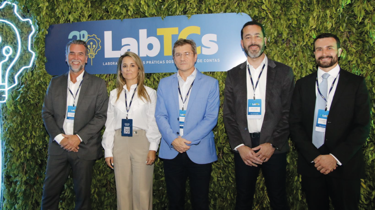 FotoFoto dos representantes do TCE/SC no evento em Cuiabá. Em frente a um fundo de folhas verdes, com uma placa com o logo do LabTCs e uma luminária de lâmpada, estão, em pé, da esquerda para a direita, Marcelo Brognoli, Gissele Nunes, José Nei Ascari, Daniel Antunes e Silvio Sallum.