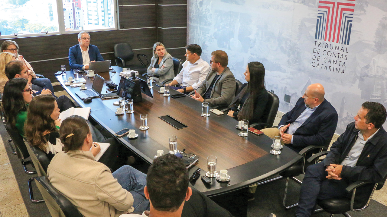 Banner horizontal com foto de reunião realizada no TCE/SC. Há várias pessoas sentadas ao redor de uma mesa, enquanto conversam. Na ponta, o conselheiro substituto Gerson dos Santos Sicca.  