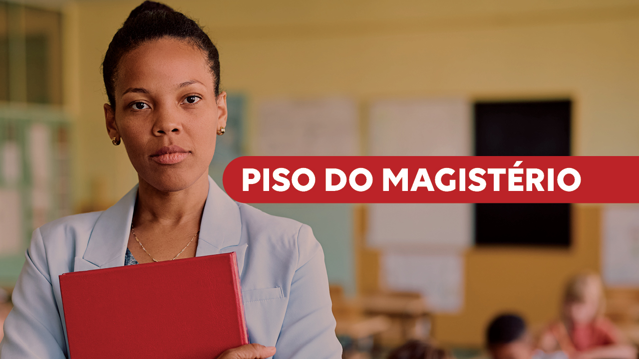 Banner horizontal com a imagem de uma professora em primeiro plano, segurando um caderno vermelho e olhando diretamente para a câmera. Ela está em uma sala de aula, com alunos e carteiras desfocados ao fundo. À direita da imagem, há uma tarja vermelha com o texto “Piso do magistério” em destaque.