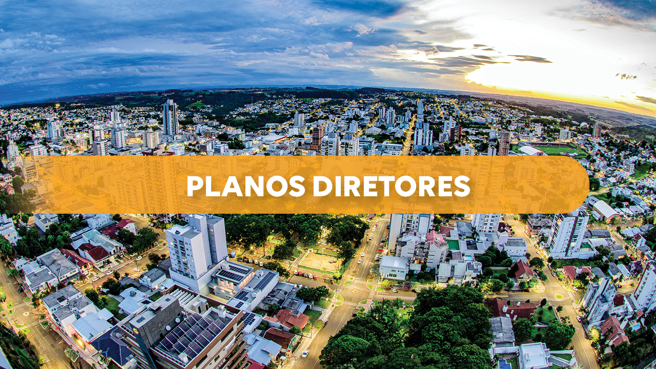 Imagem que mostra uma vista aérea de uma cidade, com prédios, ruas e áreas verdes, iluminada pela luz do entardecer ao fundo. Ao centro há uma faixa laranja com o texto “Planos Diretores”. 