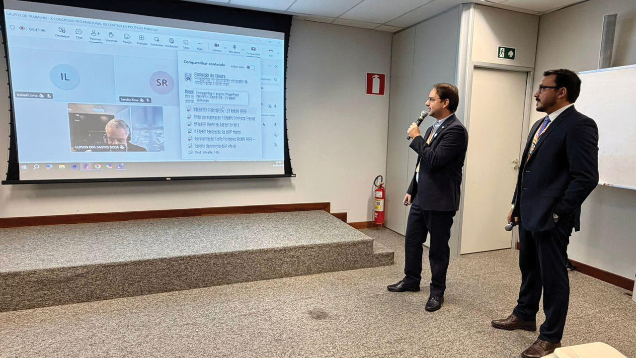Imagem horizontal que mostra os servidores Rafael Tachini de Melo e Thanderson Pereira de Sousa, lado a lado, em ambiente institucional, durante apresentação. Eles estão à direita, falando ao microfone. À esquerda, um telão projeta uma reunião, da qual participa remotamente o conselheiro substituto Gerson Sicca.