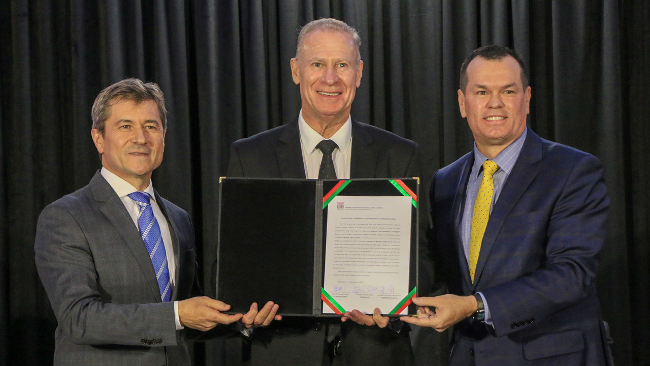 Banner horizontal com foto dos conselheiros José Nei Ascari, Herneus De Nadal e Adircélio de Moraes Ferreira Júnior no ato de posse. Eles estão em pé, um ao lado do outro, sorrindo, no palco do auditório do TCE/SC. Ambos seguram e mostram para a foto um documento assinado, dentro de uma prancheta. Atrás deles, as demais autoridades que compõem a bancada batem palmas. 