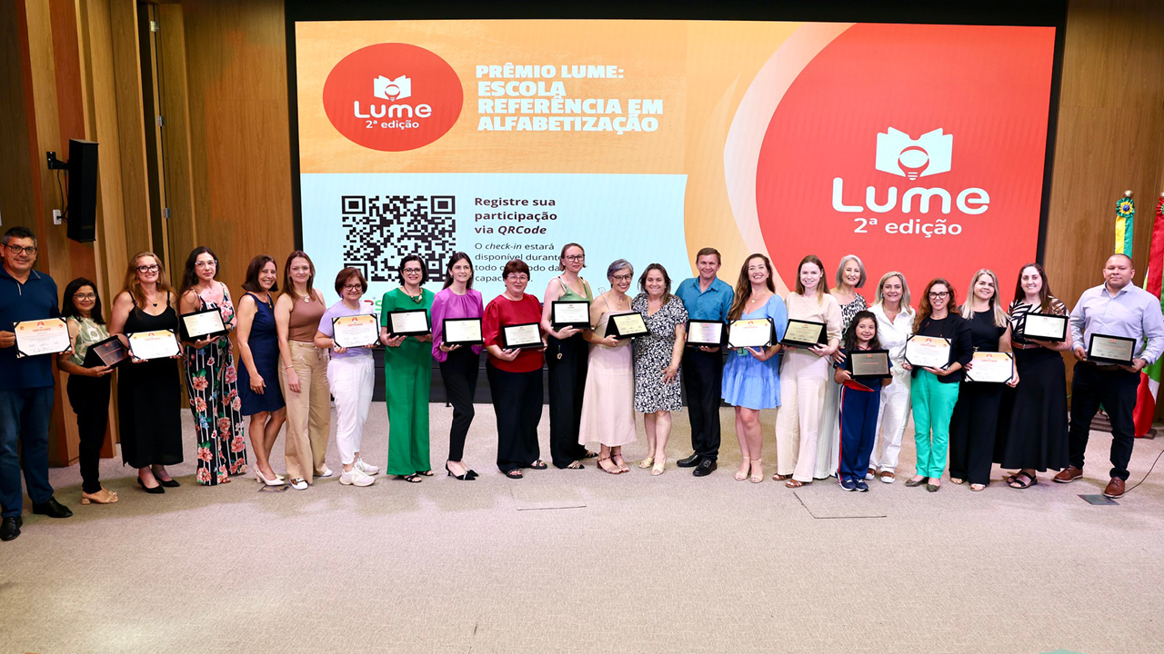 Foto que mostra representantes de todas as escolas premiadas no palco do auditório do evento. Há 23 pessoas em pé, uma ao lado da outra. A maioria segura os certificados e mostram-nos para a foto. Atrás, no telão, é projetado o logo do prêmio Lume, em tons de laranja. 