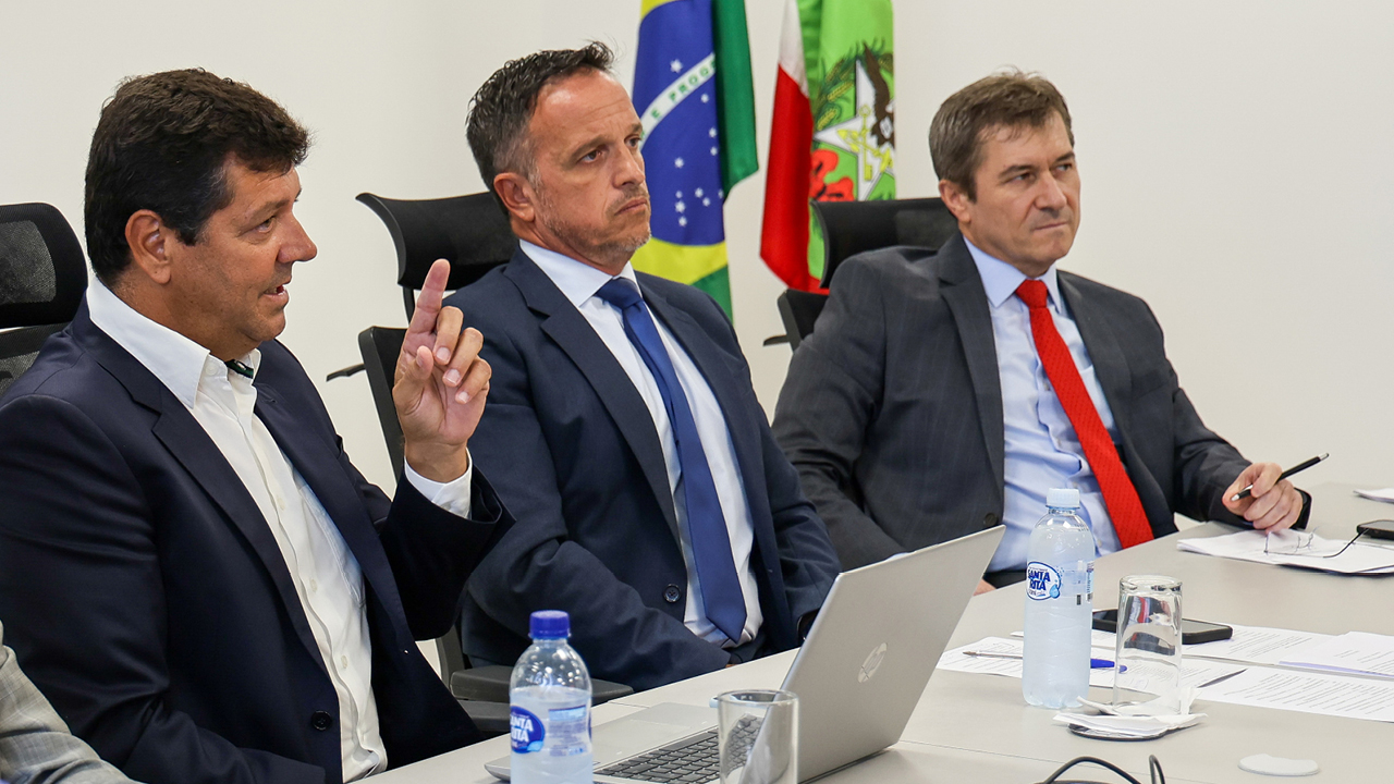 Foto, da esquerda para a direita, de Osvaldo Faria de Oliveira, de Paulo Antonio Locatelli e do conselheiro José Nei Ascari. Eles estão sentados diante de uma mesa, durante o encontro, enquanto conversam. Todos vestem terno. Sobre a mesa, há computadores, papéis, canetas e copos. Ao fundo, as bandeiras do Brasil e de Santa Catarina.