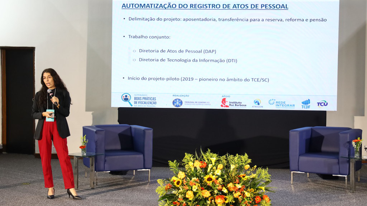 Imagem mostra uma mulher que veste calça vermelha e casaco preto em um palco, do lado esquerdo de um painel onde estão projetadas algumas frases.