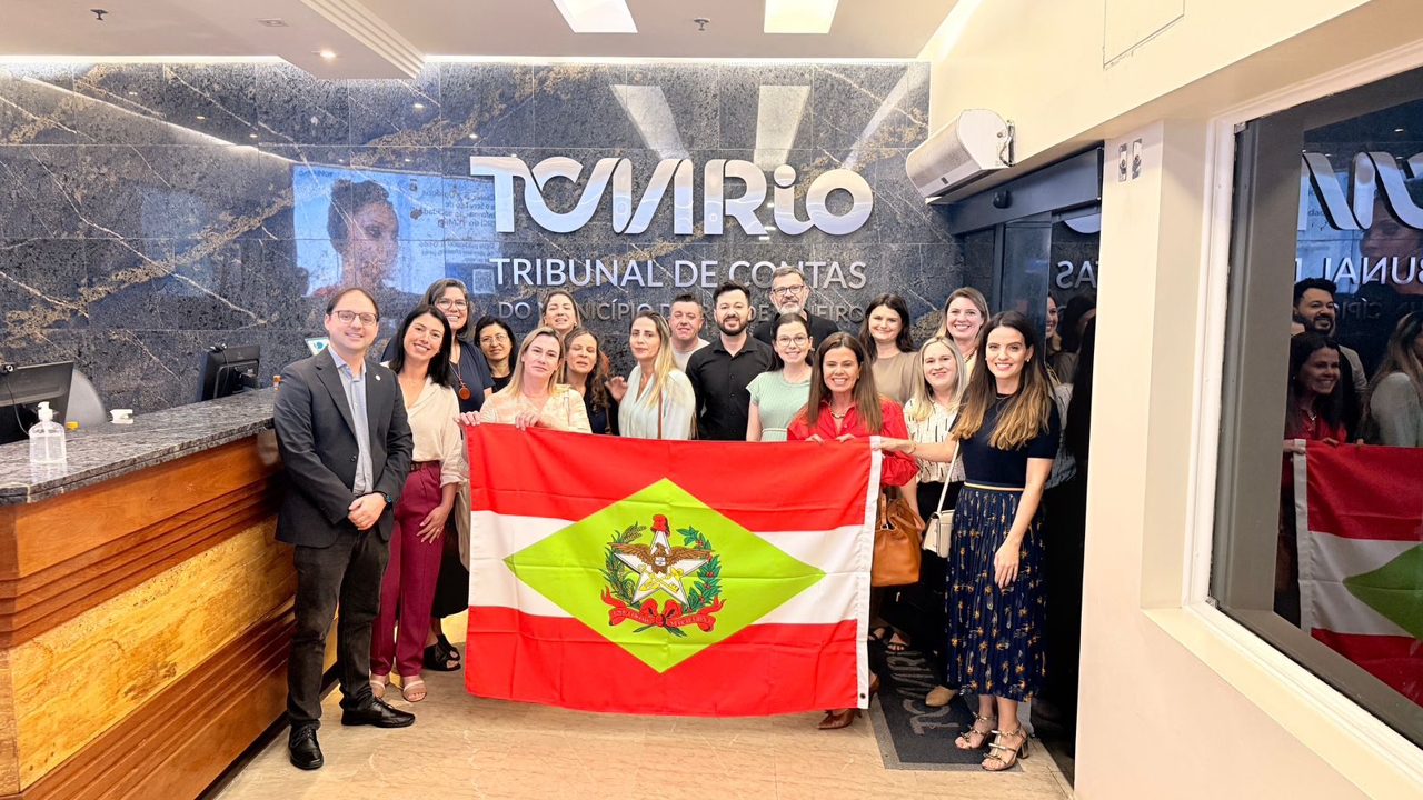 Banner horizontal com foto da comitiva catarinense segurando uma bandeira de Santa Catarina em ambiente interno no TCMRio. Entre os presentes, diretoras das escolas vencedoras e servidores do TCE/SC