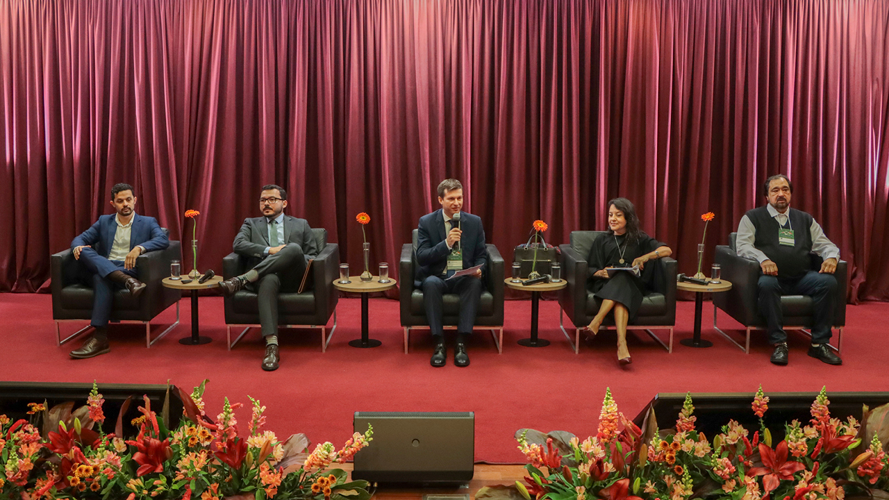 Foto mostra cinco pessoas sentadas em cadeiras em um palco. Quatro são homens e há uma mulher. Flores fazem uma espécie de moldura em primeiro plano.