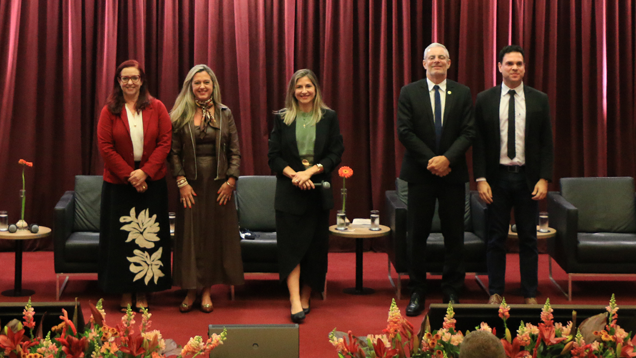 Banner horizontal com foto posada dos cinco palestrantes no palco do auditório bordô do TCE/SC. Entre os presentes, o conselheiro substituto Gerson Sicca, a diretora executiva do Icon, Marina de Miranda, a advogada Caroline Bitencourt, a presidente do IPDA, Adriana Schier, e o procurador Janriê Reck. Eles estão em pé. O cenário é composto por tapete e cortinas bordôs, além de flores decorativas. 