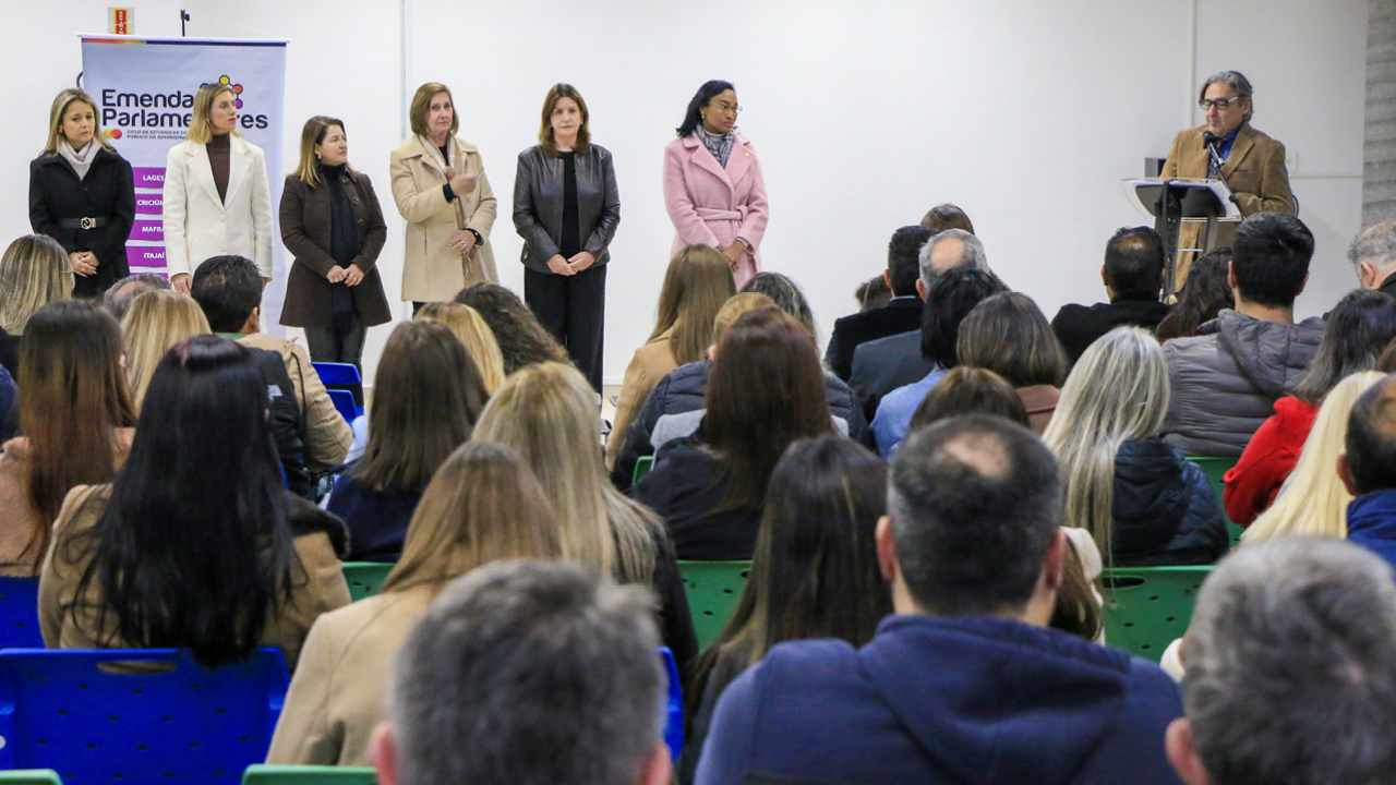 Imagem mostraum grupo de pessoas em um palco, com público em primeiro plano. À direita, há um homem falando em um microfone, atrás de um púlpito. À esquerda aparecem seis mulheres