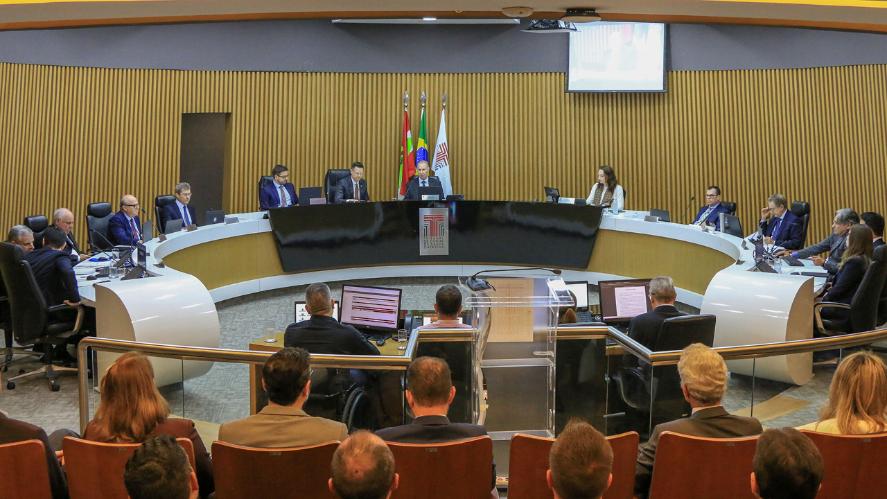 Banner horizontal com foto do plenário do TCE/SC durante sessão de apreciação das contas. Ao centro, conselheiros e conselheiros substitutos estão posicionados em uma grande mesa semicircular. No centro da composição, está a mesa diretiva da sessão, conduzida pelo presidente Herneus De Nadal e pelo relator do processo, conselheiro Luiz Roberto Herbst. Também participam da sessão procuradores MPTC/SC e representantes do governo estadual, entre eles o secretário da Fazenda, Cleverson Siewert. 