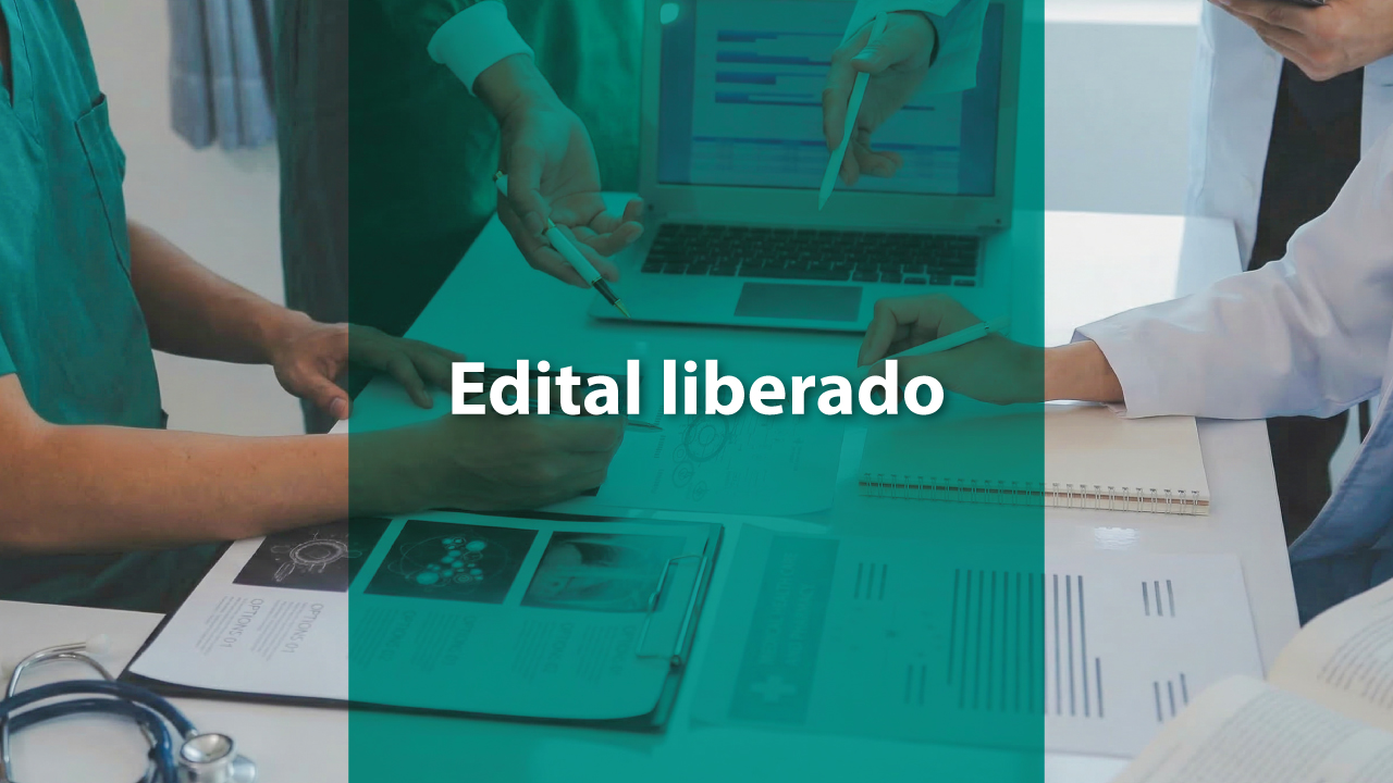 Banner horizontal com foto de profissionais da área da saúde reunidos em torno de uma mesa, analisando documentos. Sobre a imagem há uma faixa translúcida em tom de verde no centro, com o texto “Edital liberado” em destaque. Na mesa aparecem papéis, pranchetas, um notebook e um estetoscópio. 