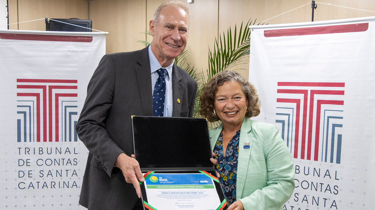 Banner horizontal com foto do presidente do TCE/SC, conselheiro Herneus De Nadal, ao lado da secretária nacional de Integridade, Patrícia Alvares de Azevedo Oliveira. Eles seguram o selo de apoiador institucional do Programa Time Brasil, entregue ao conselheiro Herneus. Ambos sorriem e estão em pé, em frente a dois banners com logo do TCE/SC.