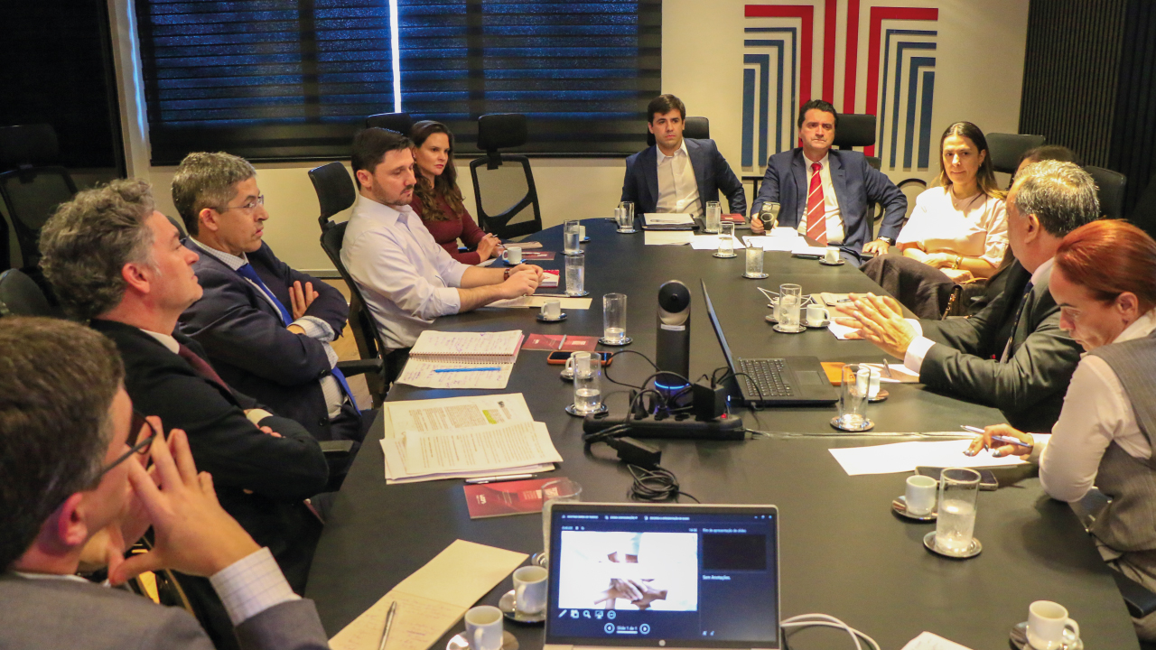 Banner horizontal com foto de várias pessoas reunidas em torno de uma mesa, enquanto conversam. Entre os presentes, o coordenador, Cassio Severo, integrantes do TCE/SC e representantes do Município de Biguaçu e da Beneficência Camiliana do Sul. Sobre a mesa, há papéis, copos de água e itens de trabalho. Ao fundo, na parede, plotagem com logo do TCE/SC.