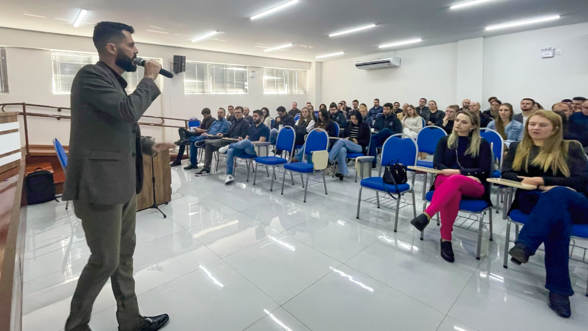 Imagem mostra à esquerda, em primeiro plano, um homem ao microfone. Ao fundo, aparecem dezenas de pessoas sentadas em cadeiras azuis, como em uma sala de aula. O teto e o chão são brancos