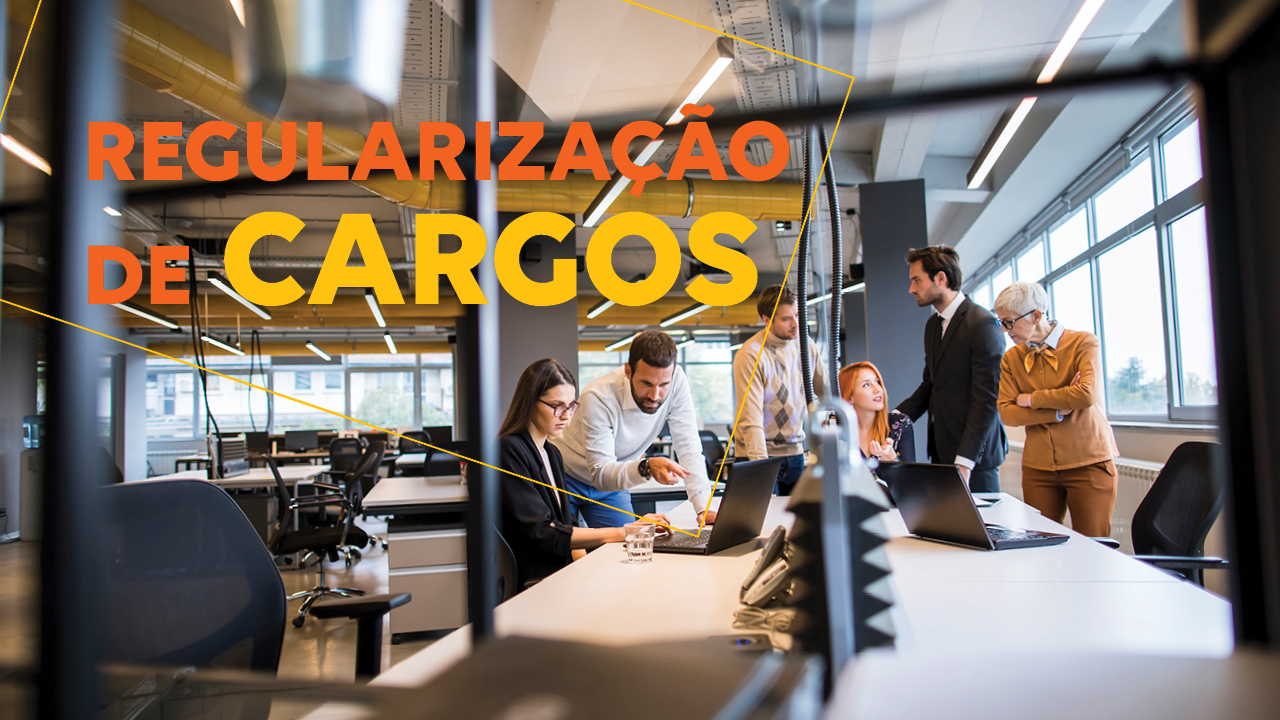 Banner horizontal com foto de um ambiente corporativo moderno, com mesas de trabalho, computadores e amplas janelas ao fundo. No centro da cena, um grupo de profissionais participa de uma reunião informal, conversando e analisando informações em notebooks. Em destaque, sobre a imagem, aparece o texto “Regularização de cargos”. 