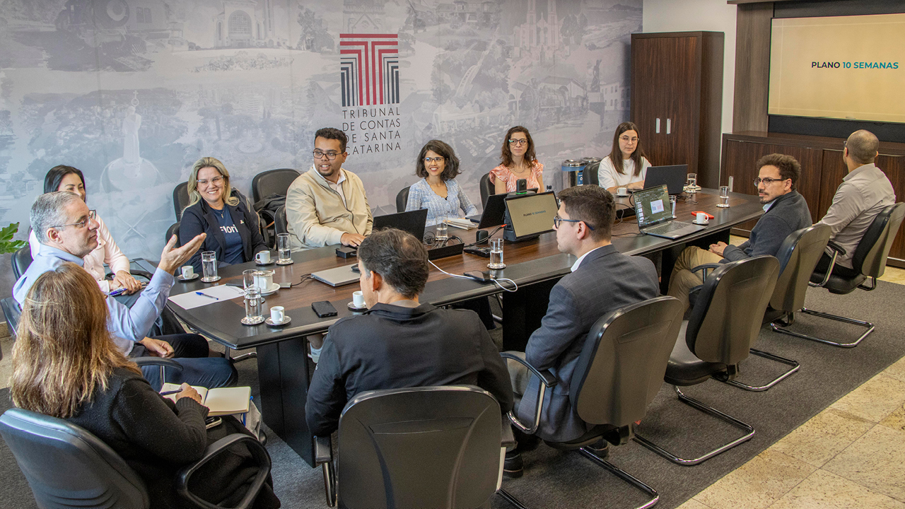 Foto horizontal de sala de reunião. Ao centro, uma mesa retangular, com doze pessoas sentadas ao redor. Na ponta da mesa, à esquerda da foto, está o conselheiro substituto do TCE/SC Gerson dos Santos Sicca. A parede ao fundo é cinza e no meio existe uma logomarca do TCE/SC. 