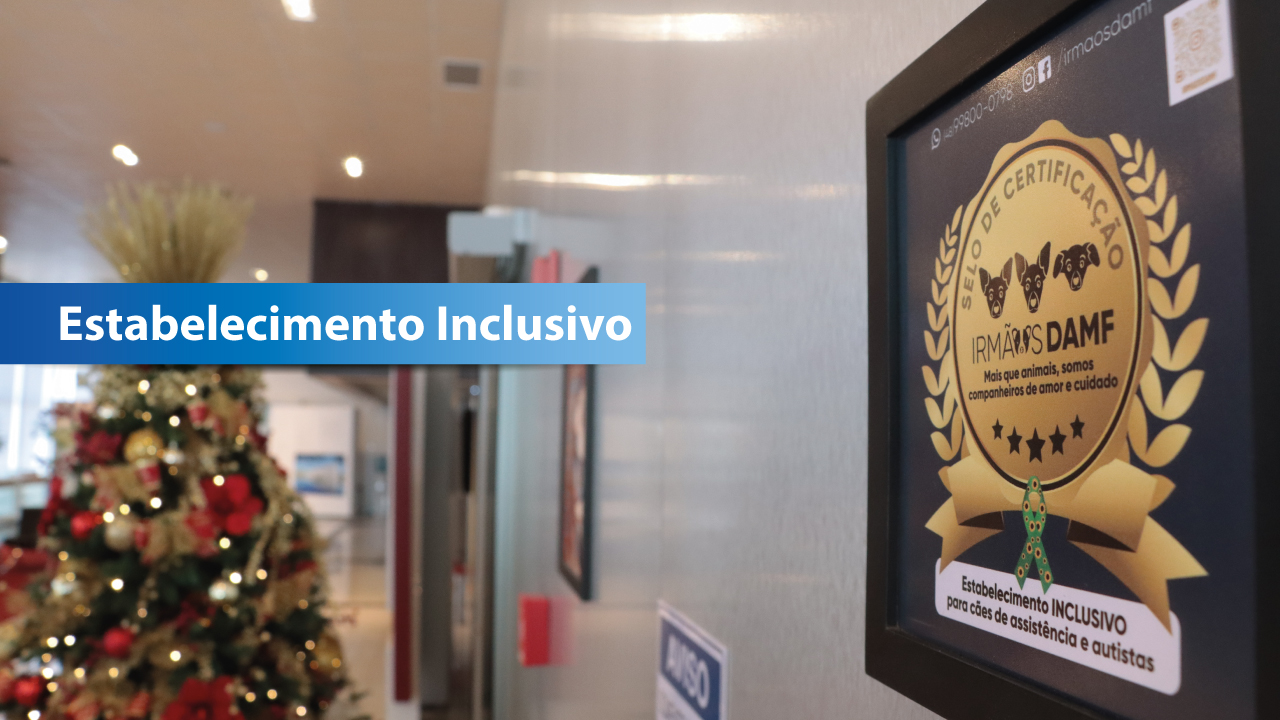 Banner horizontal que mostra uma placa colada na parede do hall de entrada do TCE/SC. Nela, o selo de "Estabelecimento inclusivo para cães de assistência e autistas". Ela tem cores preta e dourada. Ao fundo, é possível ver a árvore de Natal montada. Sobre a foto, em tarja azul, o texto "Estabelecimento inclusivo".