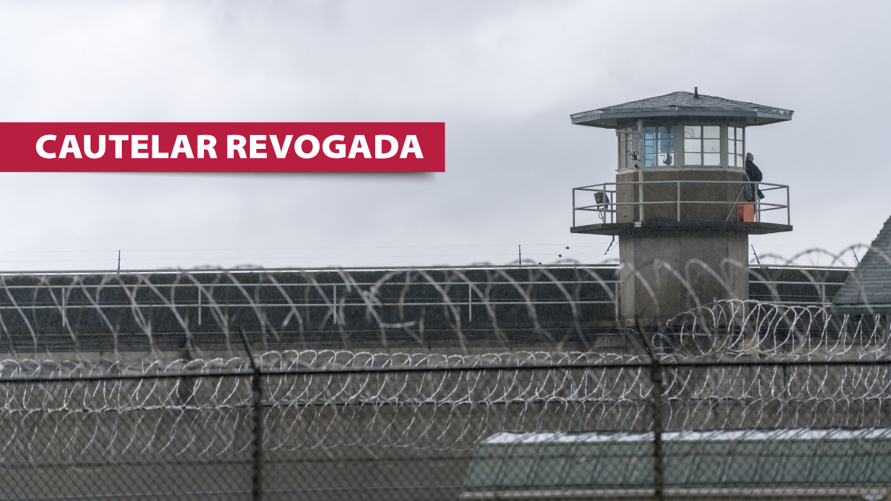 Imagem mostra o muro de uma penitenciária e uma torre de vigilância. À esquerda, escrito em branco sobre tarja vermelha, está escrito “cautelar revogada”