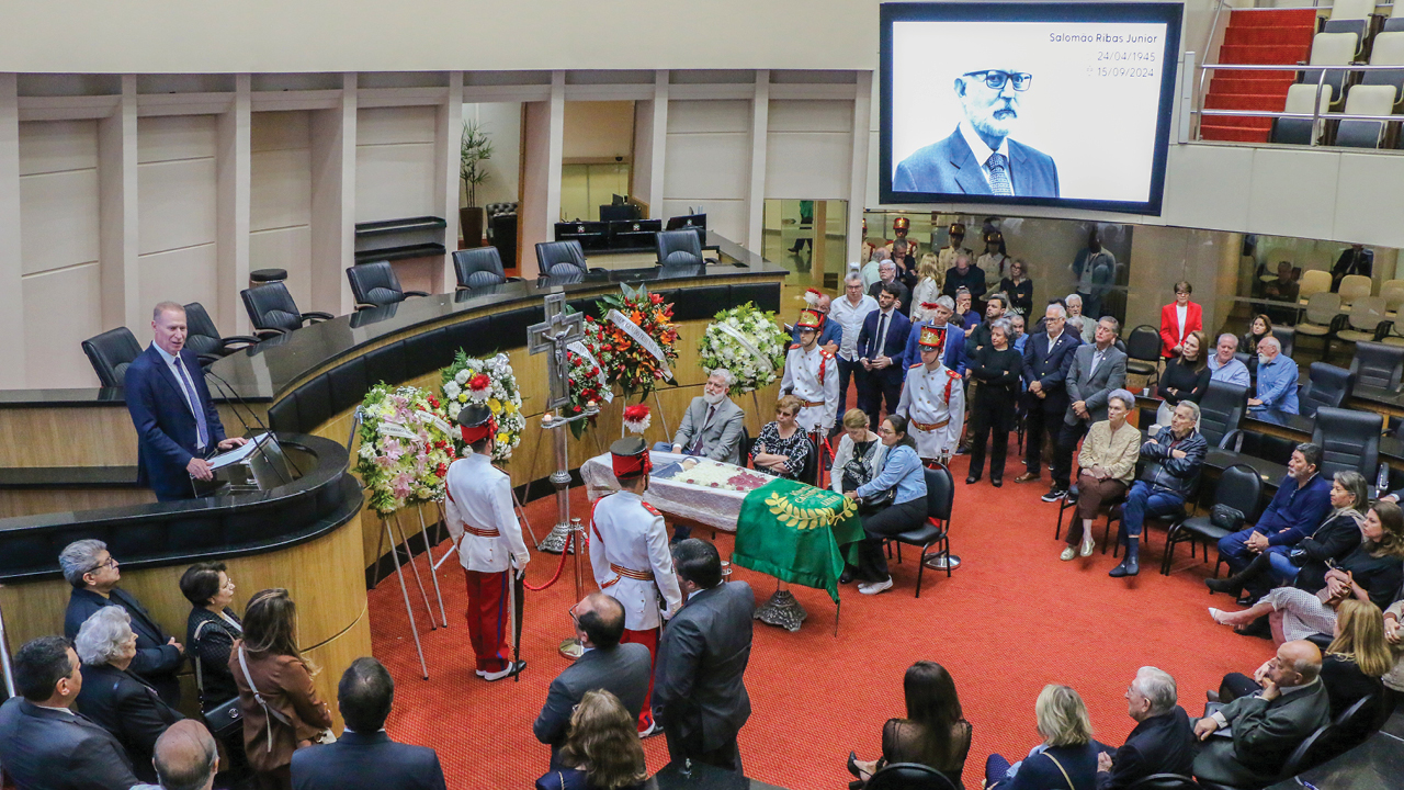 Foto do plenário da Assembleia onde ocorre o velório do conselheiro Salomão. Na Tribuna, o presidente do TCE/SC Herneus De Nadal discursa. À frente da tribuna, no centro do plenário, o caixão, ao redor do qual estão sentados familiares, guardas fardados em postura de respeito, e diversas coroas de flores. Ao redor do plenário estão sentadas várias pessoas ouvindo o discurso. No alto, no telão, a imagem do conselheiro Salomão. 