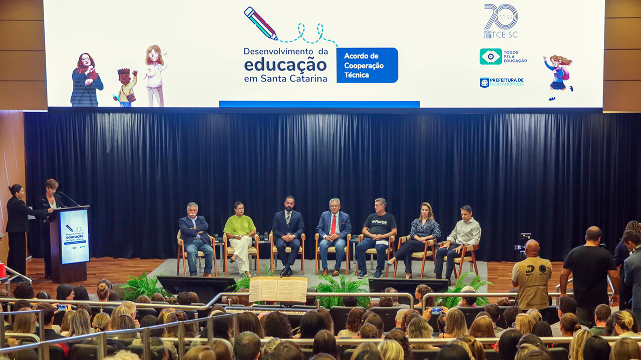 Foto horizontal dos participantes da assinatura do acordo, no palco do auditório do TCE/SC. Há sete autoridades, sentadas uma ao lado da outra. Entre os presentes, o conselheiro do TCE/SC Aderson Flores, o prefeito de Florianópolis, Topázio Neto, e a presidente executiva da Todos Pela Educação, Priscila Cruz. À frente deles, o público que assistia à solenidade, de costas, sentado em poltronas do auditório. 