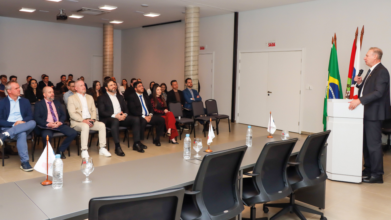Banner horizontal com foto do presidente Herneus De Nadal, à direita, discursando no púlpito do auditório. À esquerda, aparece a plateia, composta por várias pessoas sentadas lado a lado em poltronas. 