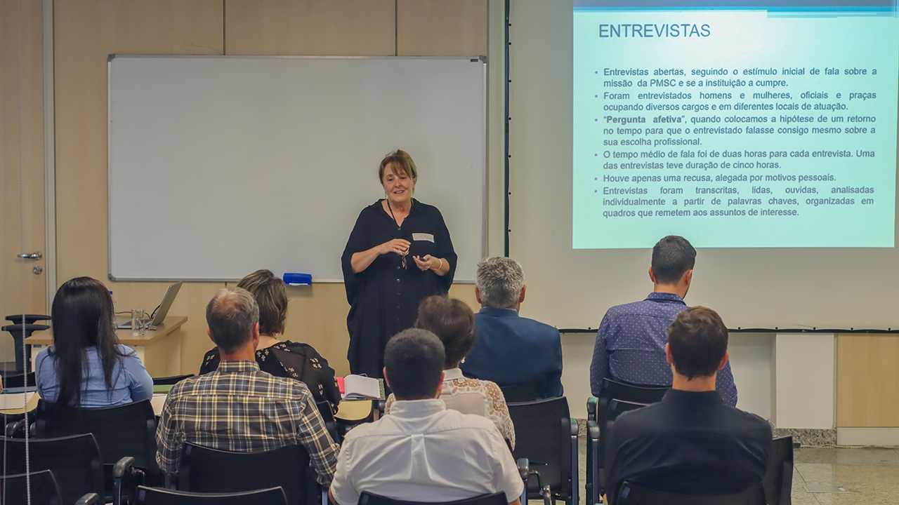   Foto de uma das pesquisadoras apresentando trabalho aos auditores do TCE/SC. Ela está em pé, de frente para o público. Eles estão de costas, sentados em carteiras enfileiradas. Atrás dela, há um telão que projeta a apresentação. 