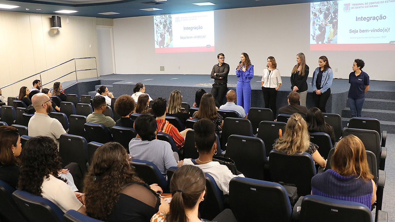 Foto do auditório azul do TCE/SC durante a recepção dos novos residentes. Em primeiro plano, de costas para a foto, estão os ingressantes, sentados em poltronas azuis. À sua frente, ao fundo da imagem, há seis pessoas em pé, fazendo uma apresentação. Iamara, da DGP, fala ao microfone.